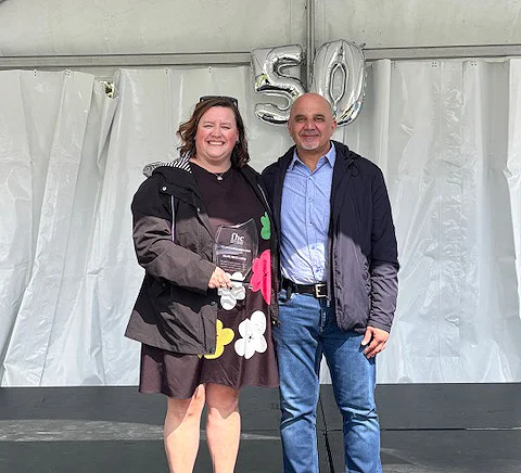 A woman holding an award stands beside a man under white tent drapes. They both smile as silver balloons form "50" in the background, conveying celebration.