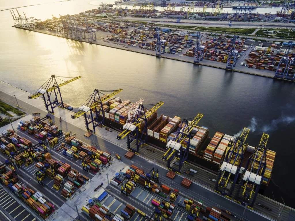 Aerial view of a bustling port at sunset with large cranes loading colorful shipping containers onto docked cargo ships, conveying industrial activity.