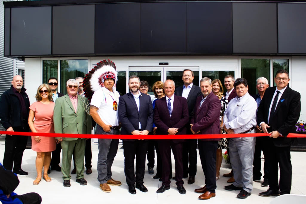 Ribbon cutting ceremony with 17 people all lined up behind the ribbon. A man in the centre of the photo and slightly left is wearing a traditional headdress and white t-shirt