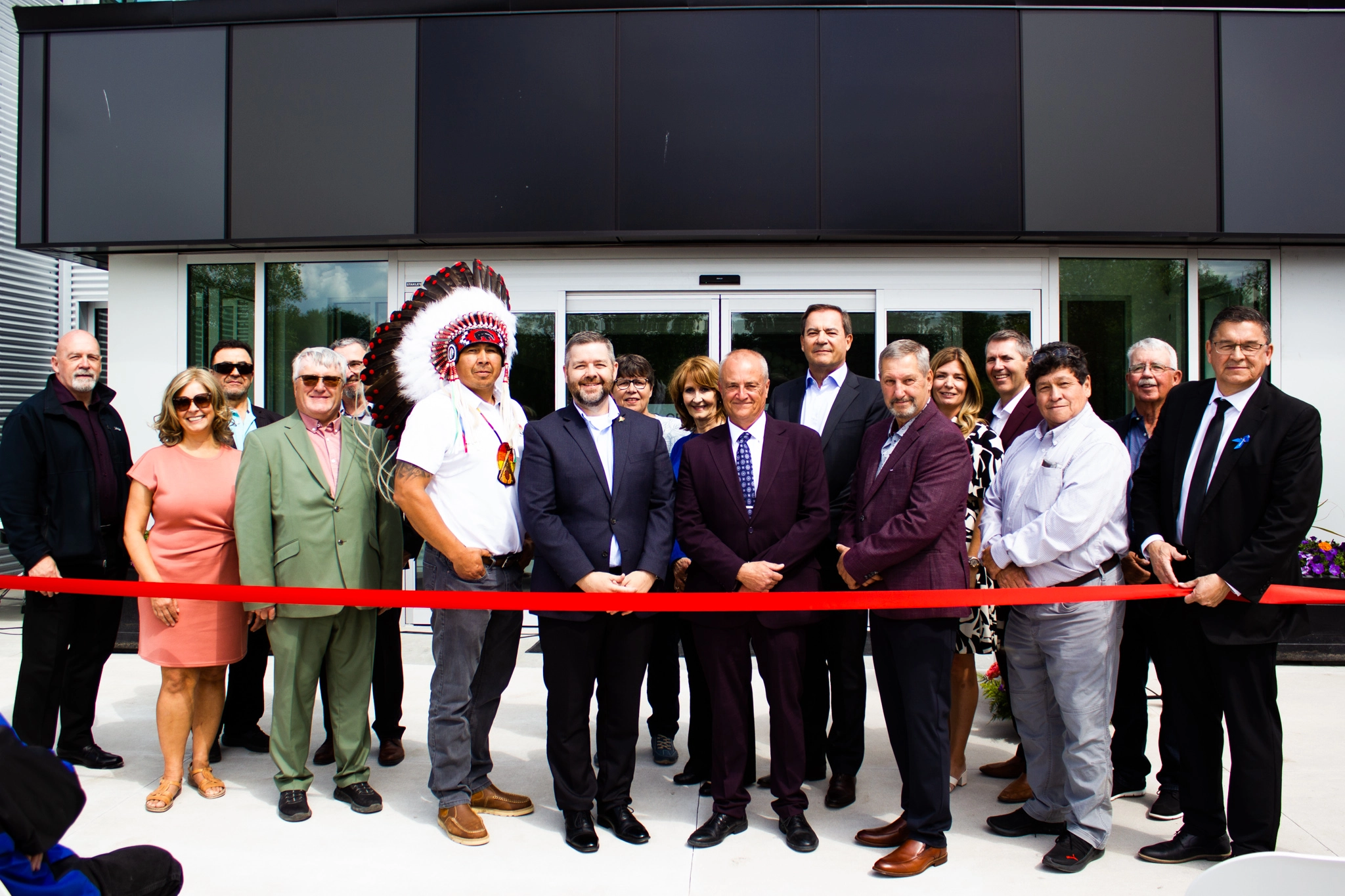 Ribbon cutting ceremony with 17 people all lined up behind the ribbon. A man in the centre of the photo and slightly left is wearing a traditional headdress and white t-shirt